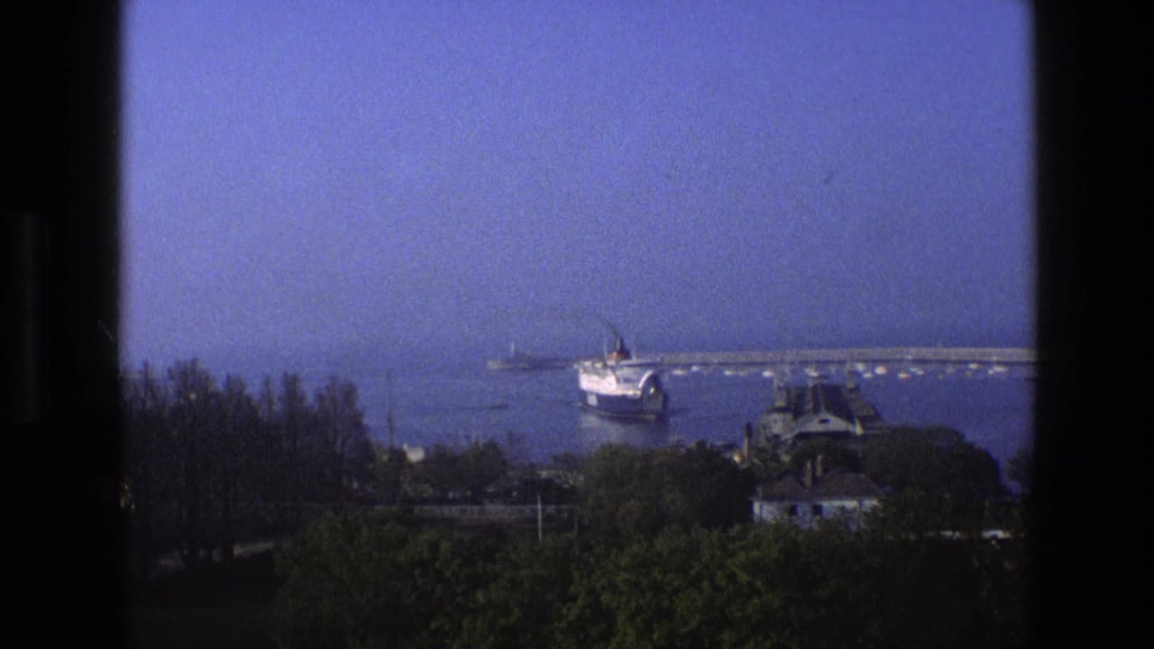 1969 England home-movie view of a passenger ship approaching a harbor, framed by dark window edges with trees below.