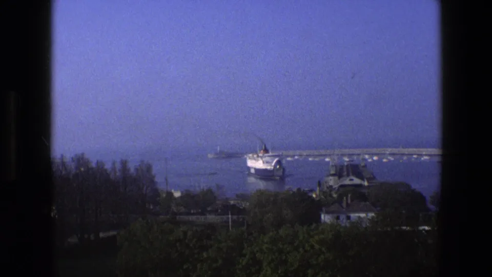 1969 England home-movie view of a passenger ship approaching a harbor, framed by dark window edges with trees below.