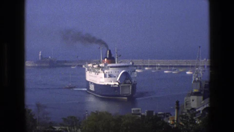 Closer 1969 England 8mm view of a passenger ferry entering port, its dark funnel releasing smoke near small boats and a breakwater.