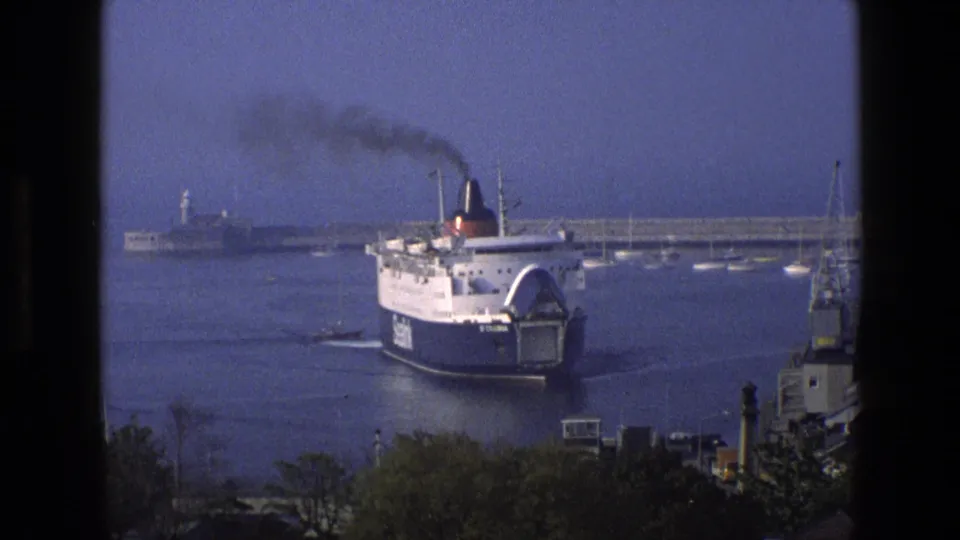 A passenger ferry turns within the harbor in 1969 England footage, with black smoke, moored boats, trees, and dock equipment visible.