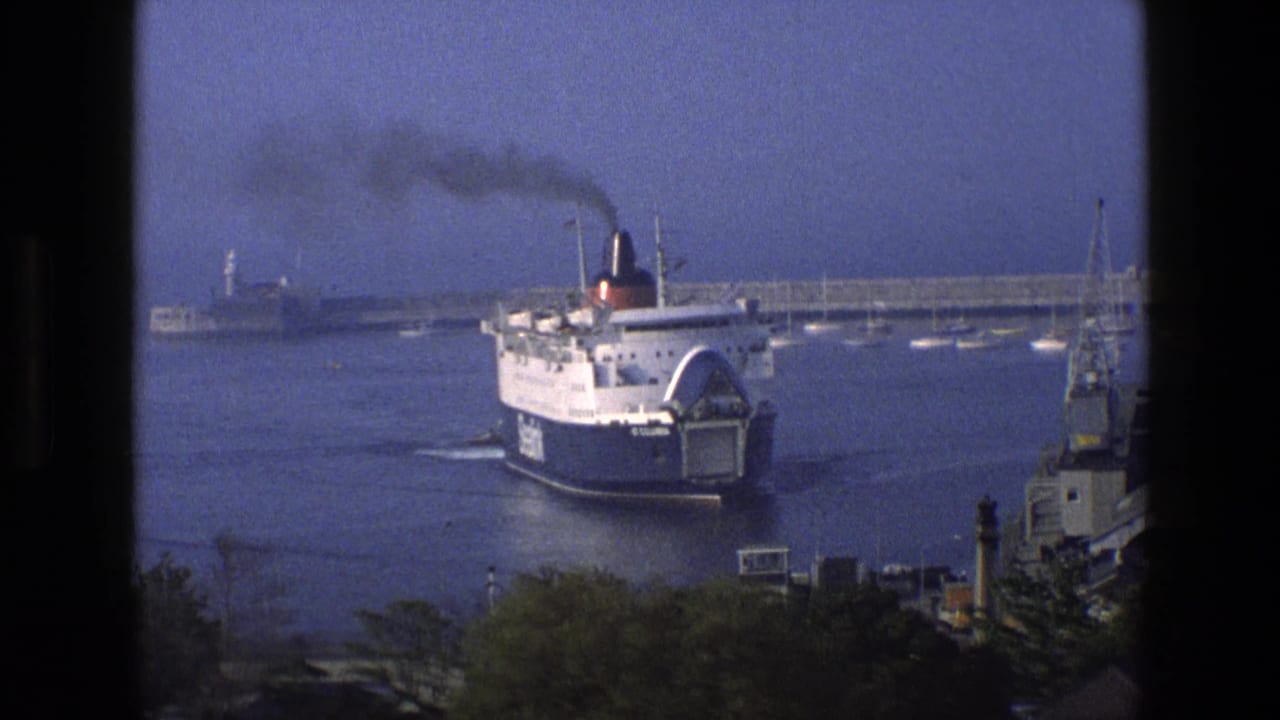 White and blue ferry moving through calm harbor water near a breakwater.