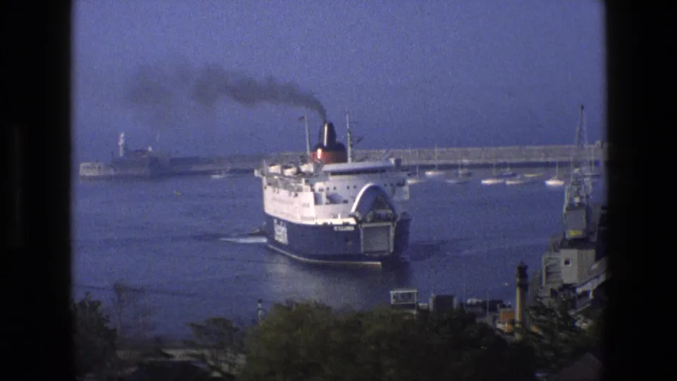 Wide 8mm view of the St Columbia ferry entering an English harbor, with smoke, small boats, and a lighthouse visible behind it.