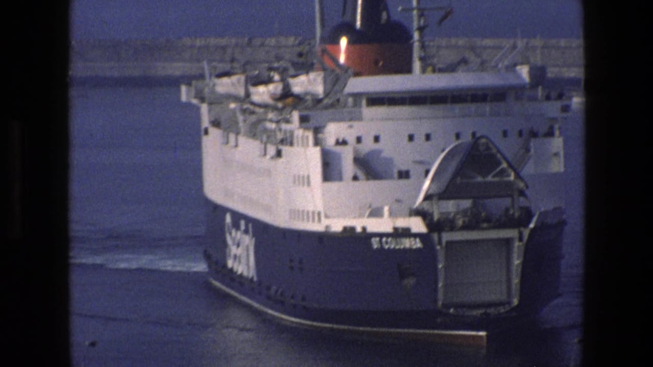 Ferry stern and ramp seen on calm water near a harbor breakwater.