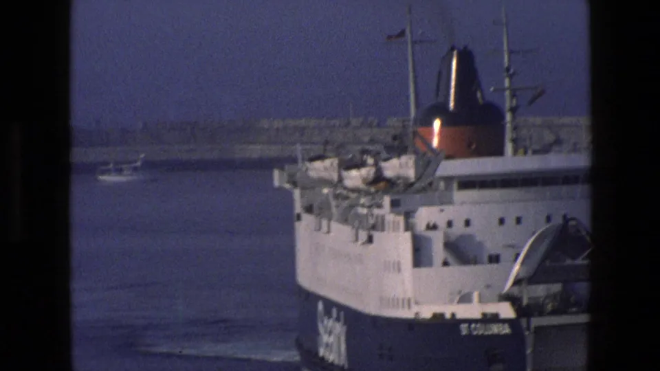 1969 England 8mm frame showing a large passenger or ferry vessel at harbor, with water and distant shoreline visible.