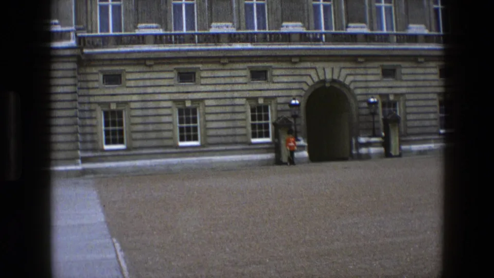 1969 England home-movie frame with a ceremonial guard standing still beside the palace entrance and sentry box.