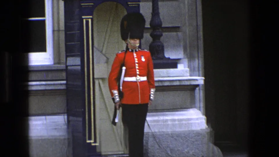 Closer 1969 England home-movie view of a British ceremonial guard in red tunic and tall black hat holding a rifle at his post.