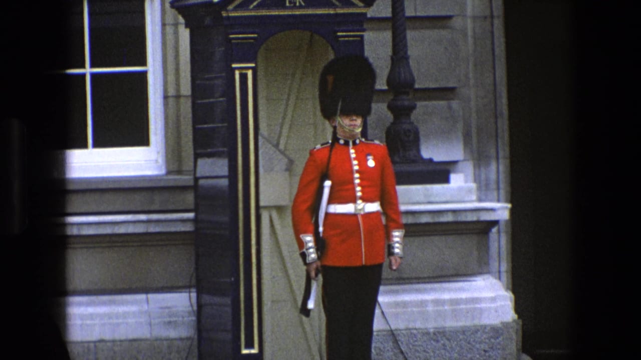 1969 England home-movie frame of a British ceremonial guard in red tunic and bearskin hat beside an ER-marked sentry box.