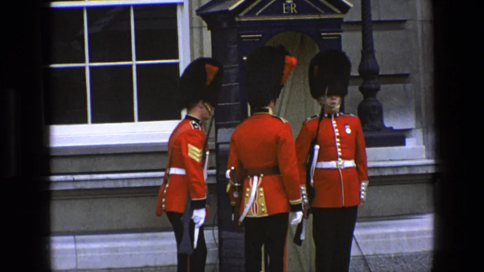 1969 England home-movie frame of British ceremonial guards in red tunics grouped near a sentry box outside a stone building.