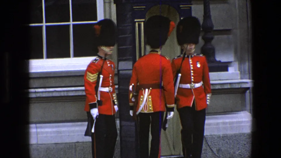 1969 England frame of British soldiers in red ceremonial dress and bearskin hats standing by an ER-marked guard post.