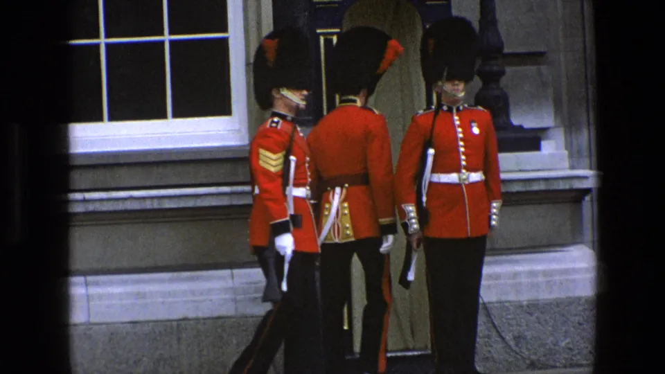 In 1969 England, guards in red tunics and bearskin hats stand at attention near a sentry box outside a grand stone facade.