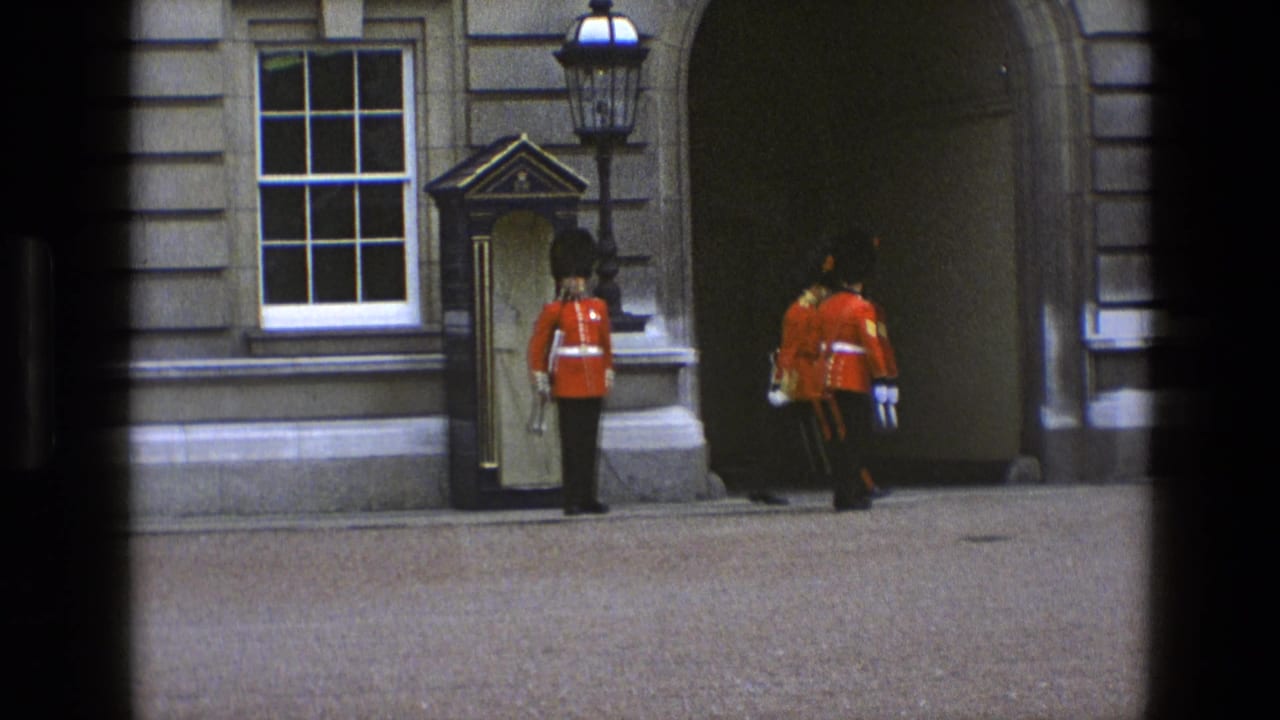 In 1969 England, one guard stands at a sentry box as several red-coated guards move toward a large stone archway.
