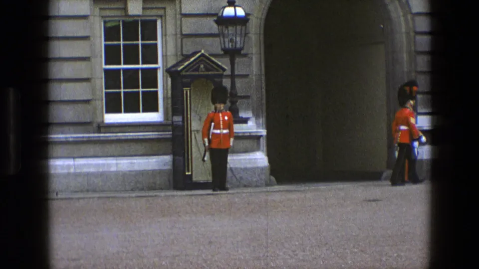 In 1969 England, a guard stands by a sentry box as another guard passes near the palace archway and stone frontage.