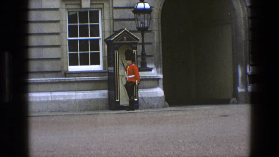 In 1969 England, a single red-coated guard stands near a sentry box beside a large stone archway at the palace frontage.