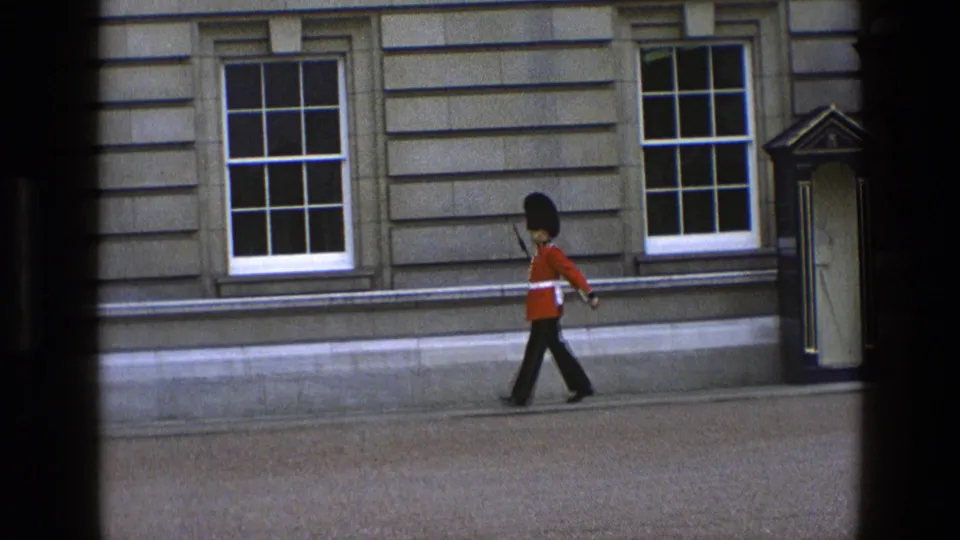 1969 England 8mm frame showing a ceremonial guard in red tunic marching along a stone building exterior.