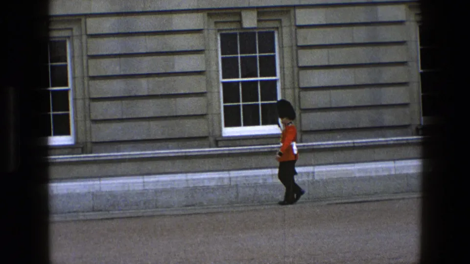 1969 England 8mm frame of a red-coated guard continuing patrol outside a formal stone building, seen through dark foreground framing.