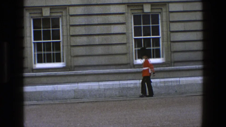 In 1969 England footage, a ceremonial guard in red tunic and tall black hat walks past sash windows on a stone facade.