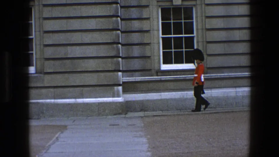 The guard in red tunic and black bearskin-style hat moves past a projecting stone corner and sash window in England footage from 1969.