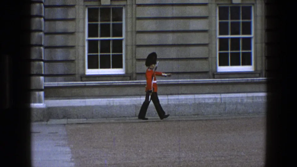 The guard continues marching in ceremonial uniform along the stone facade, with the camera view divided by dark vertical foreground edges.