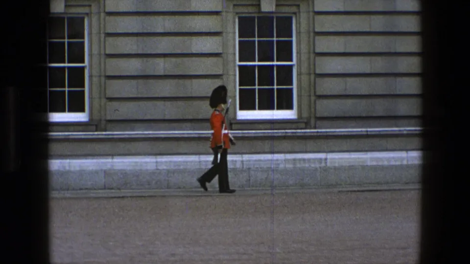 A ceremonial guard in red tunic and black bearskin-style hat moves farther along the stone courtyard wall in 1960s England footage.