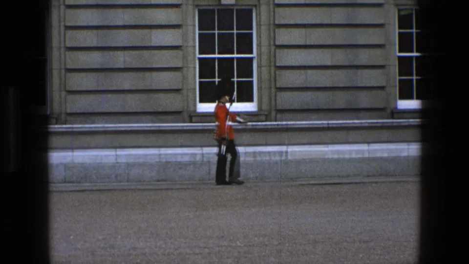 In 1969 England footage, a guard in red tunic and tall black hat marches beside a stone facade with sash windows.
