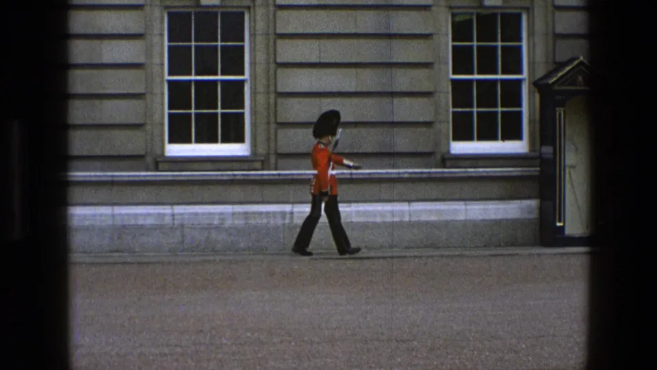 A British ceremonial guard appears in profile, marching toward a sentry box against a stone building facade.