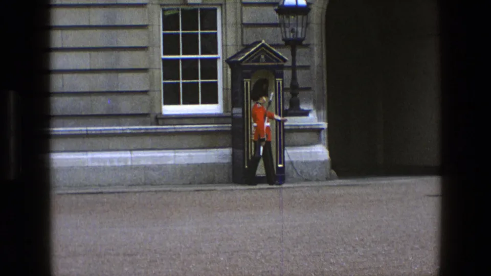 In 1969 England footage, a ceremonial guard in a red tunic and black hat stands near a sentry box, window, and lamppost.