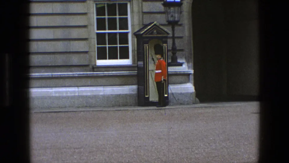 The guard faces forward near the sentry box, holding a rifle upright against the stone exterior in archival 8mm footage.