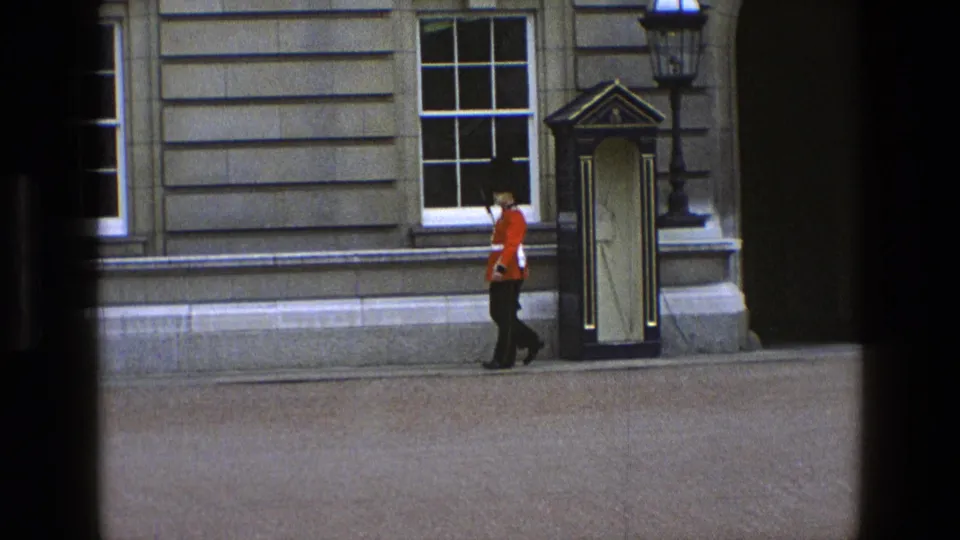 The guard walks past the sentry box and stone facade, continuing the ceremonial movement in the 1969 England home-movie clip.
