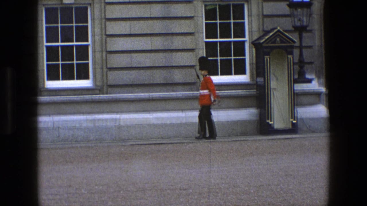 In England 1969, a ceremonial guard in red tunic and black hat walks past a sentry box along a stone facade.