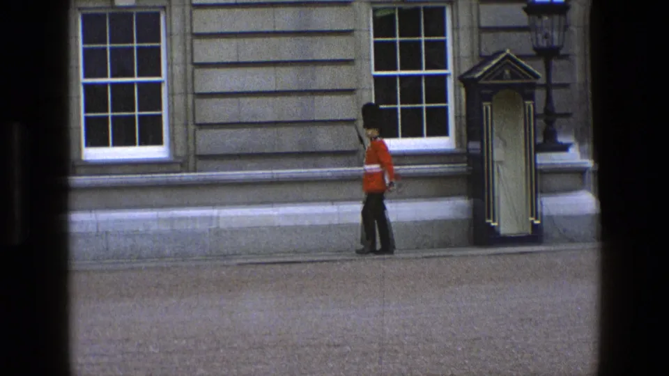 In England 1969, a ceremonial guard in red tunic and black hat walks past a sentry box along a stone facade.