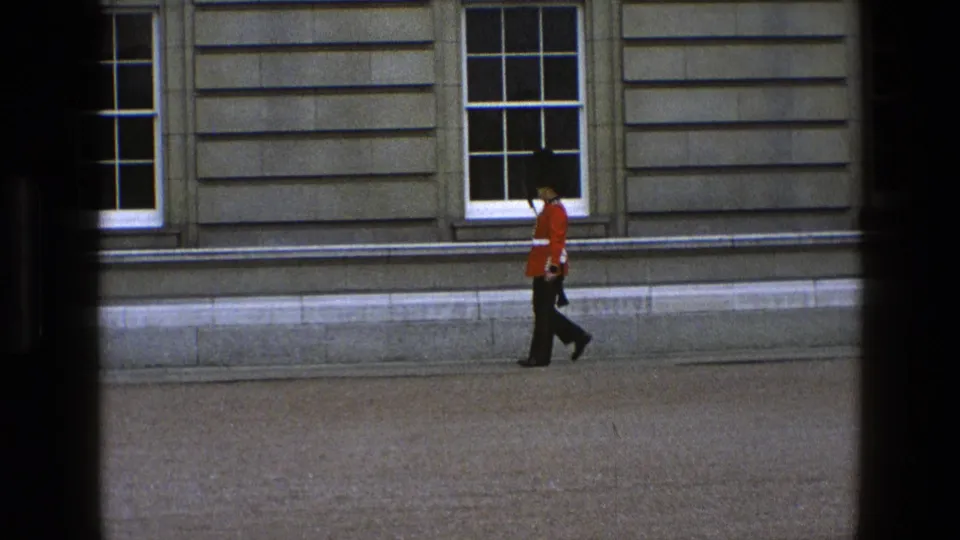 In England 1969, the guard moves past the lower part of a stone facade, with tall windows behind him.