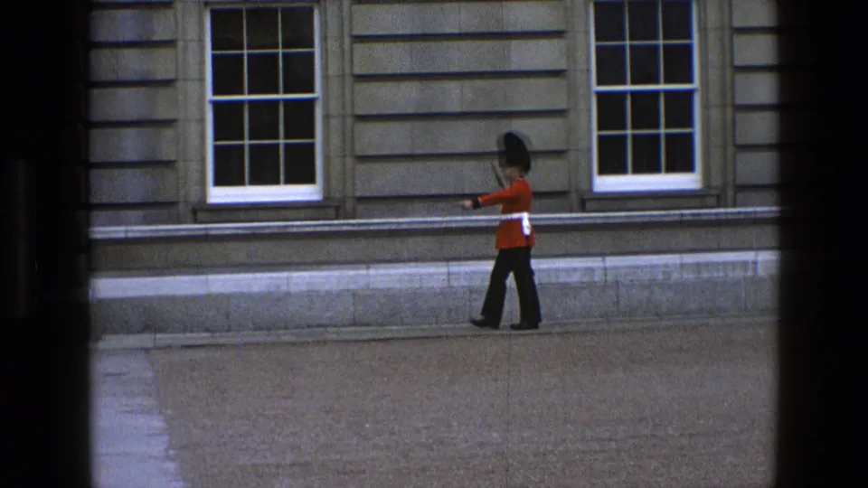 In England 1969, the guard strides along the stone exterior with his rifle held at his side.