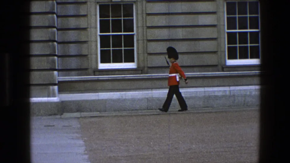 In 1969 England footage, a Queen's Guard in red tunic and tall black hat walks along a stone facade with sash windows.