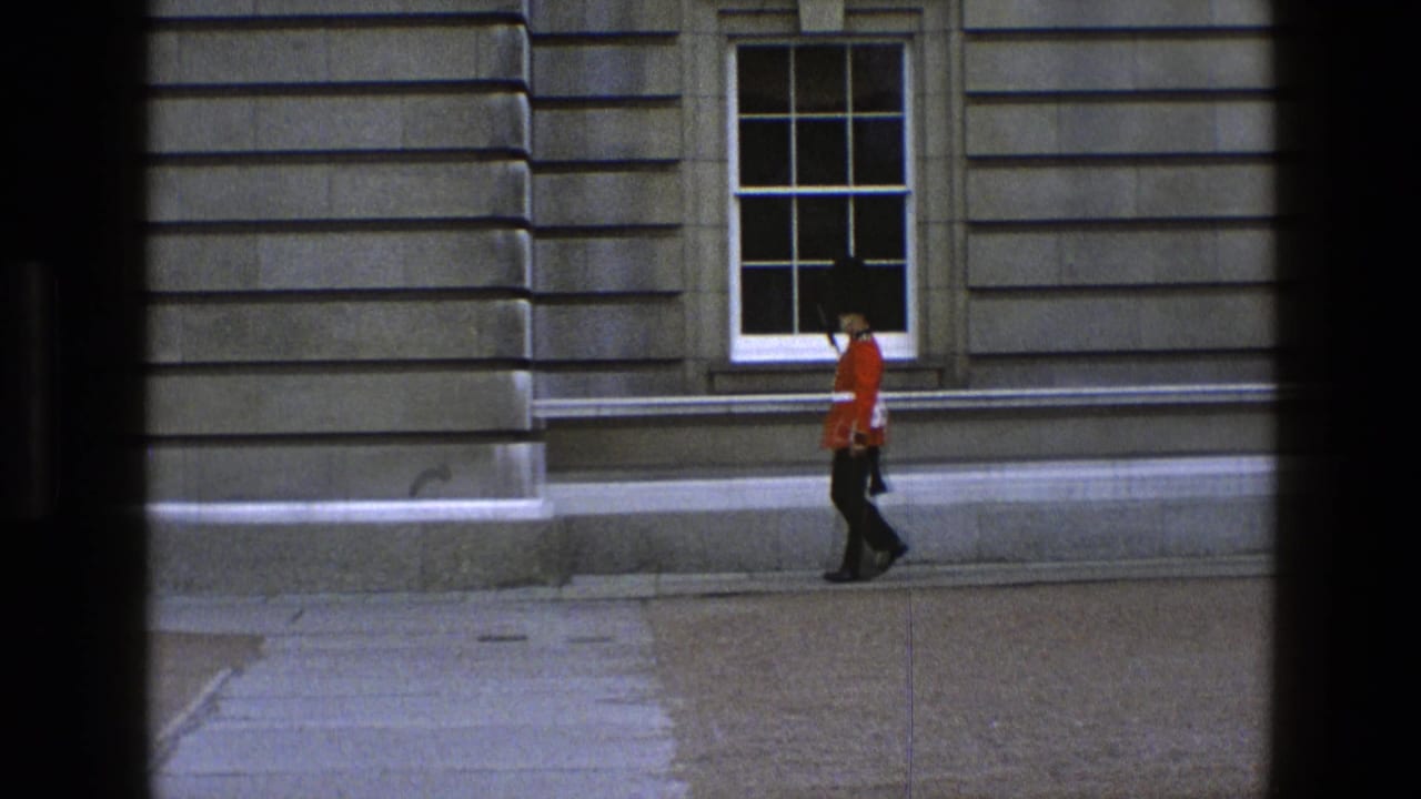 Guard in red uniform walks beside a stone wall with window behind.