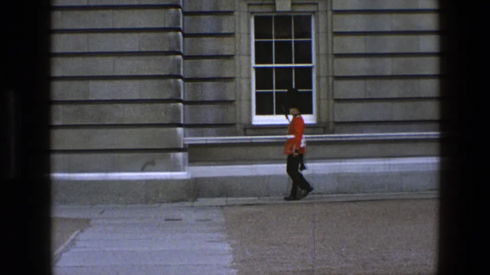 A British ceremonial guard in red uniform and black bearskin-style hat passes a large window on a stone building.