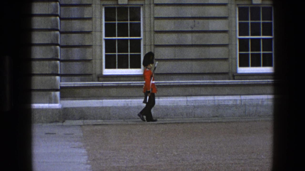 A British ceremonial guard in red uniform and black bearskin-style hat moves along a stone building facade in England, 1969.