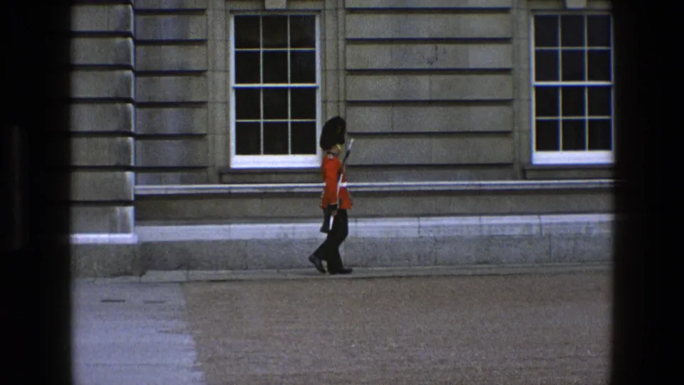 A British ceremonial guard in red uniform and black bearskin-style hat moves along a stone building facade in England, 1969.