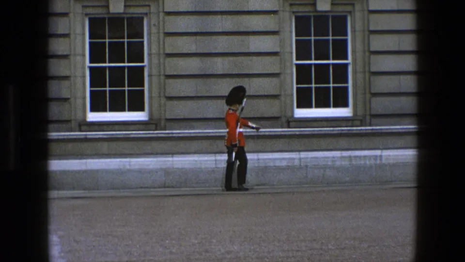The guard appears to pause or turn near the stone building, with red tunic, black hat, rifle, and window details visible.