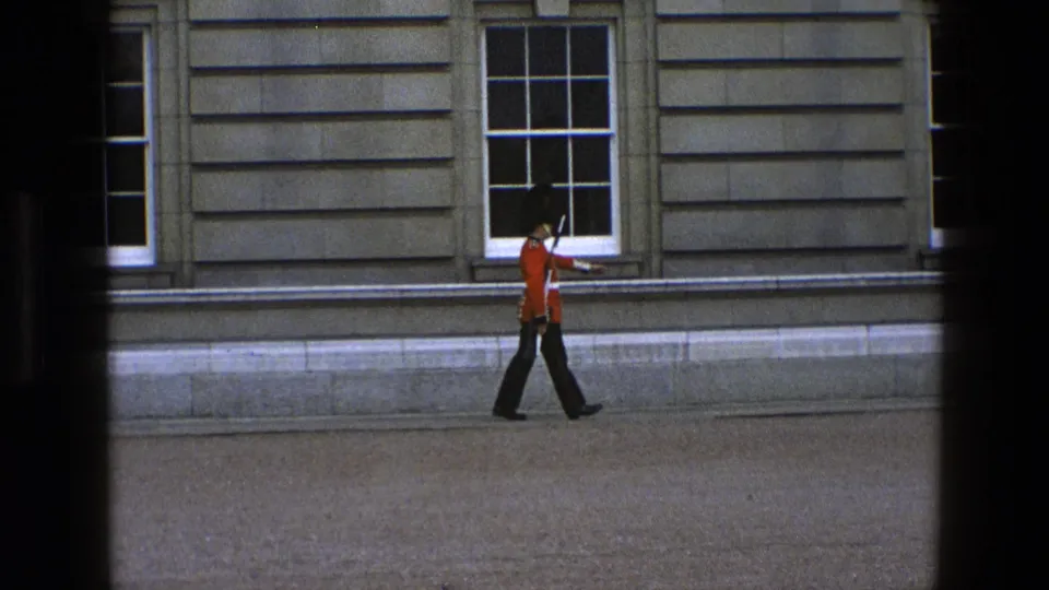 In a home-movie view, the British ceremonial guard marches across the courtyard edge with a rifle against a formal stone facade.
