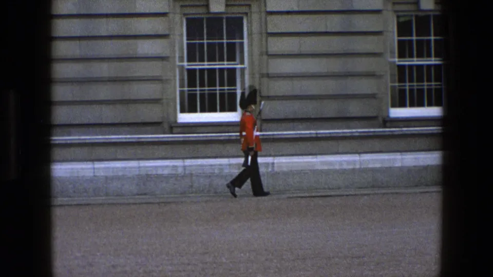 In 1969 England footage, a British ceremonial guard in red tunic and tall black hat walks past sash windows carrying a rifle.