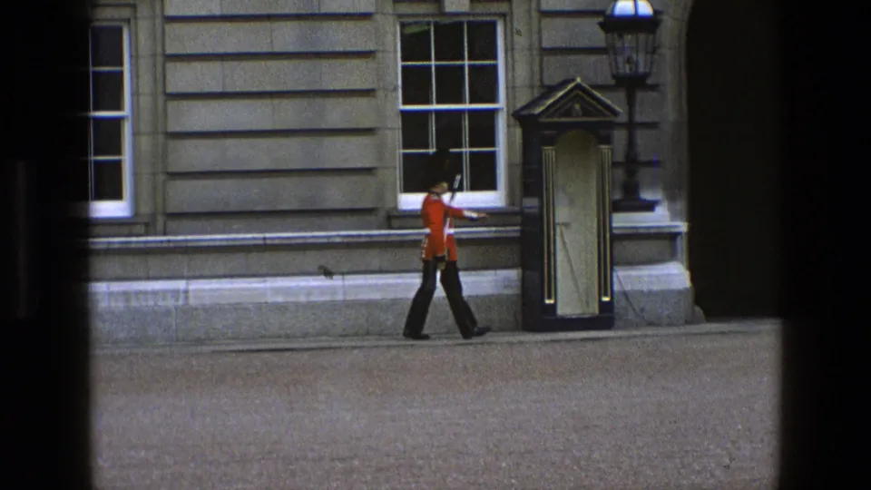 The guard continues along the stone facade toward a sentry box and ornate lamp, with the rifle held at his side.