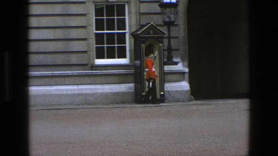 A British ceremonial guard pauses or turns near the sentry box, partially framed by the building entrance area and stone facade.