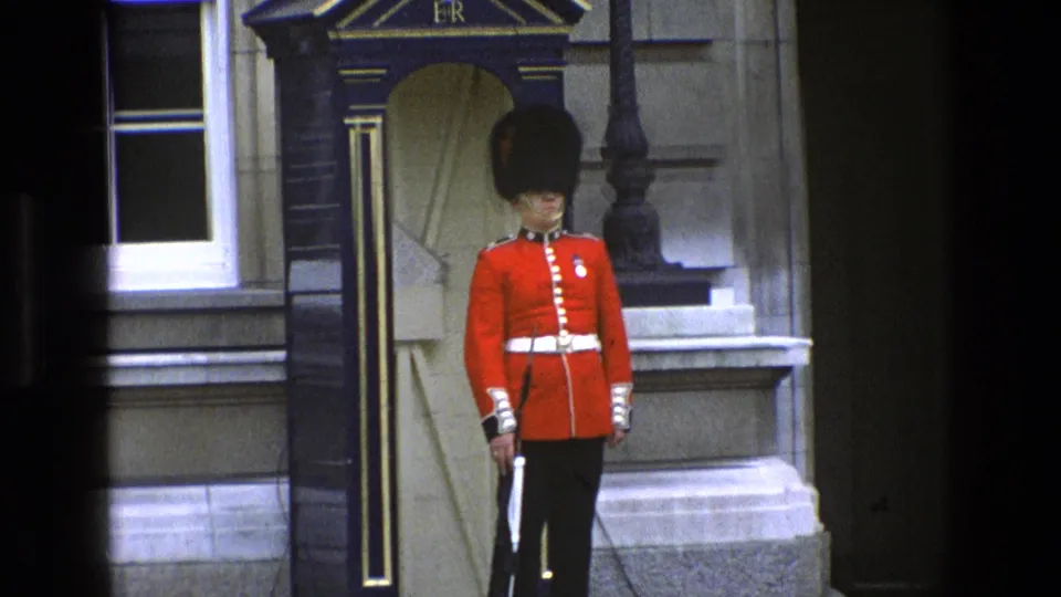 Closer 1969 England home-movie view of a royal guard in red tunic and tall black hat beside the sentry box.
