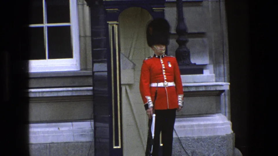 A royal guard in red tunic and tall black hat stands at attention outside a stone building in England, 1969 metadata.