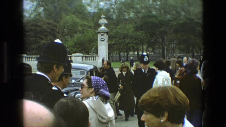 Tourists and police officers gather in an outdoor public area in England, with older cars, railings, trees, and a stone gatepost visible.