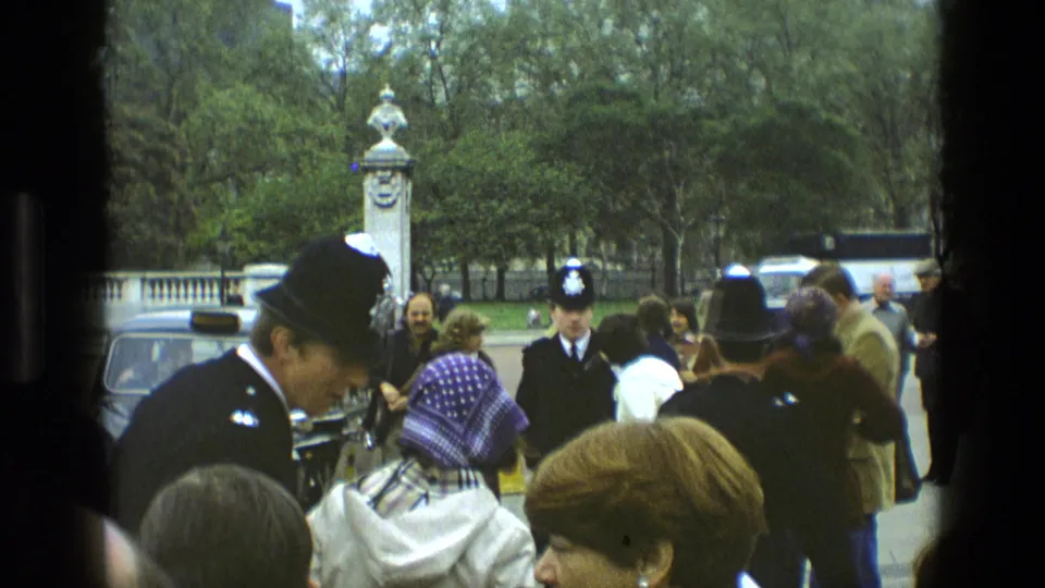 A British police officer in a helmet stands among tourists near parked cars and trees, continuing the 1969 England public crowd scene.