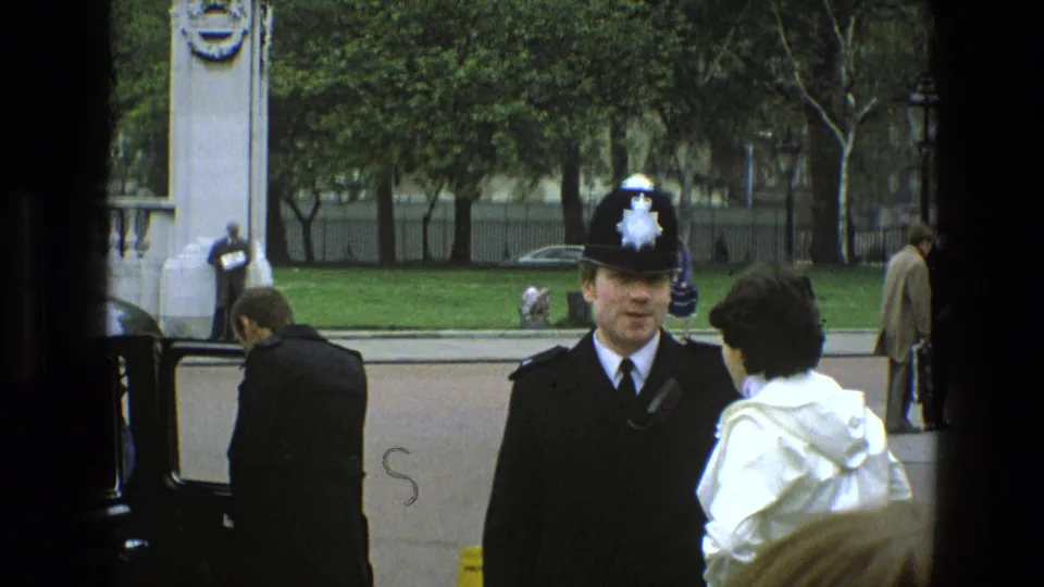 In 1969 England, a uniformed British police officer faces a person in a white coat near an open vintage car door on a tree-lined street.