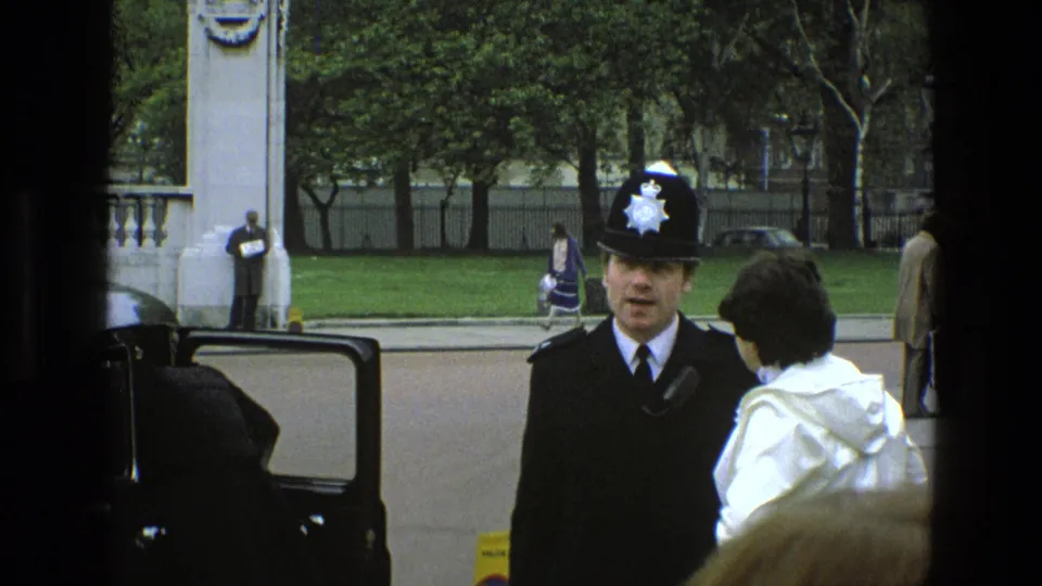 The officer continues speaking with the white-coated person as pedestrians pass behind them near stone gateposts and a green lawn in England.