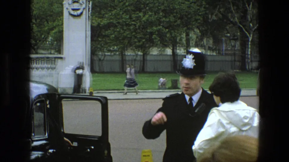 A uniformed British police officer gestures during a curbside conversation with a white-coated person, with a woman walking in the background.