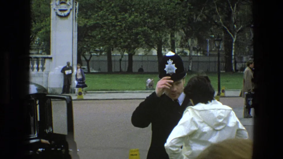 The British police officer lifts a hand to his helmet while the white-coated person remains beside him on the street in 1969 England.